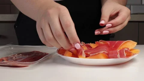Wrapping prosciutto around melon slices on white plate. Hands preparing Stock Footage 316676177