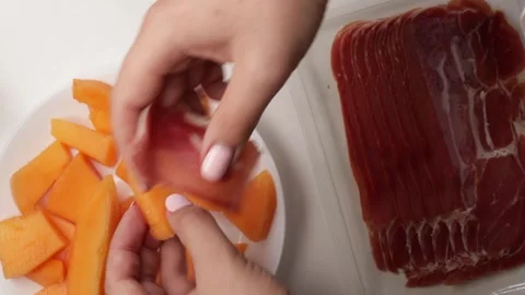 Wrapping slices of melon with jamon on white plate. Hands preparing prosciutto Stock Footage 316675582