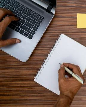 Writing on a notepad and typing on a keyboard while working on a project. A l Stock Photos