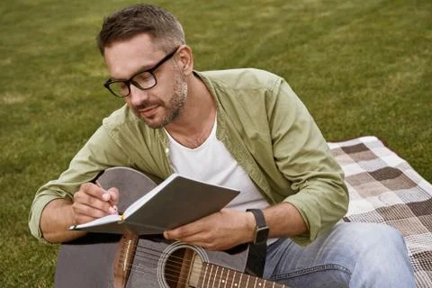 Writing notes. Young focused man wearing eyeglasses holding acoustic guitar and Stock Photos