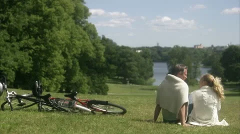 WS Rear view of couple sitting on lawn next to bikes, facing lake Stock Footage 11341124