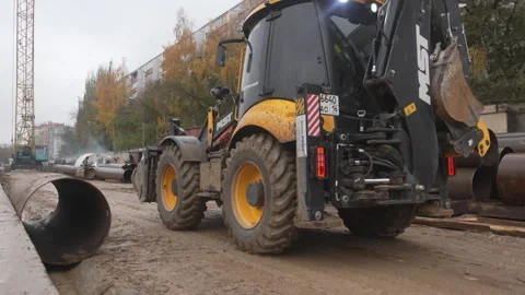Yellow backhoe drives past stacks of metal pipes in city Stock Footage 313828173