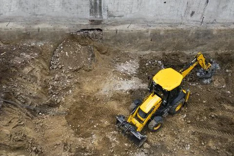 Yellow backhoe loader digging in a muddy construction pit, high angle Stock Photos