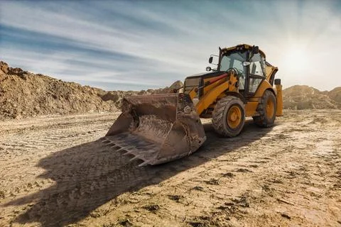 A yellow backhoe loader digs into the earth on a construction site as the sun Stock Photos