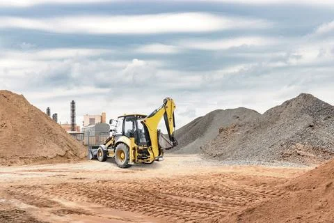 A yellow backhoe works on a construction site, digging through mounds of dirt Stock Photos