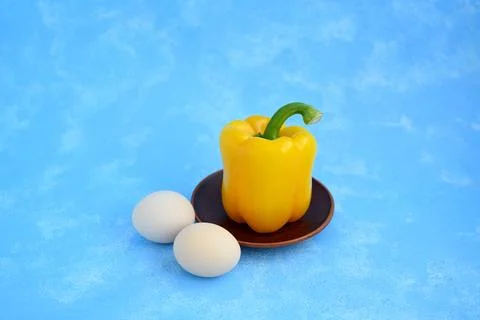 Yellow Bell Pepper with Two Eggs on a Brown Clay Plate Against Blue Background Stock Photos