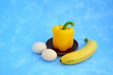 Yellow Bell Pepper with Two Eggs and a Banana on a Brown Clay Plate Against Blue Stock Photos