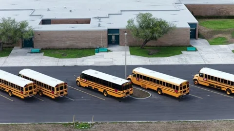 Yellow buses angle across empty asphalt lot beside low brick school building Stock Footage 340863753