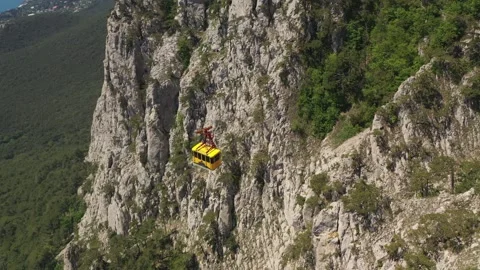 A yellow cabin of cableway going by cable along the rocky sheer wall of mount Stock Footage 165453004