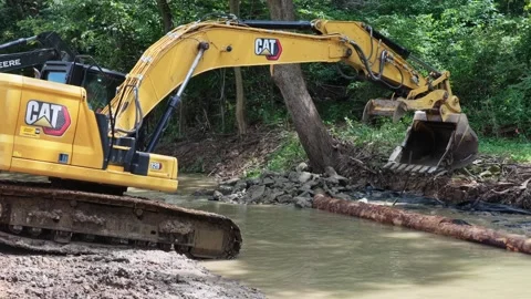 Yellow CAT excavator digs into muddy creek bed during Donegal Creek streambank Stock Footage 344564793