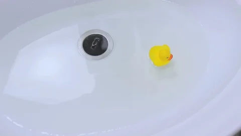 Yellow classical rubber duck floats in clean water in white ceramic bathroom sin Stock Footage 128882195