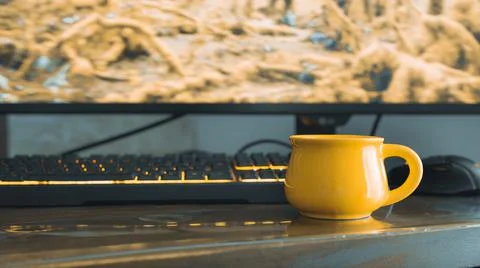Yellow coffee mug on computer desk and gaming keyboard and monitor in background Stock Photos