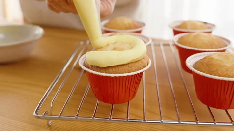Yellow cream is squeezed in a circular pattern from a pastry bag onto a cupcake Stock Footage 266476840