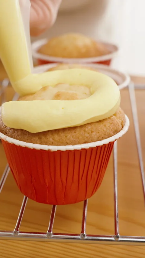 Yellow cream is squeezed in a circular pattern from a pastry bag onto a cupcake Stock Footage 266476951