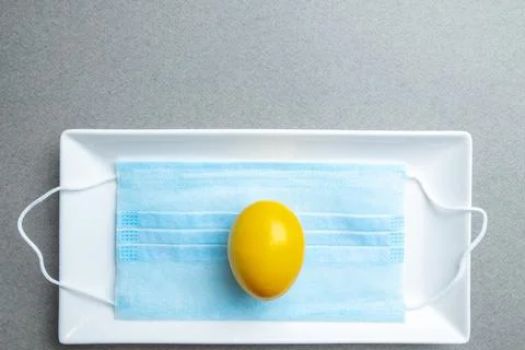 Yellow easter egg lies on a blue medical mask inside a white ceramic plate Stock Photos