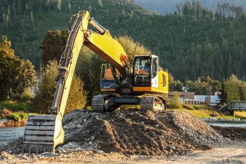 Yellow excavator (digger machine) digging stones, in river, forest in backgro Stock Photos