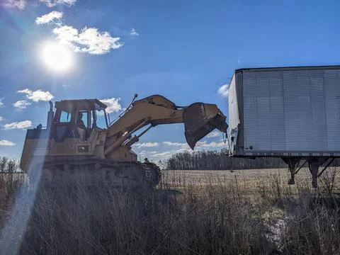 A Yellow Excavator Pulls an Abandoned Trailer in a Rural Field Under Blue Skies Stock Photos