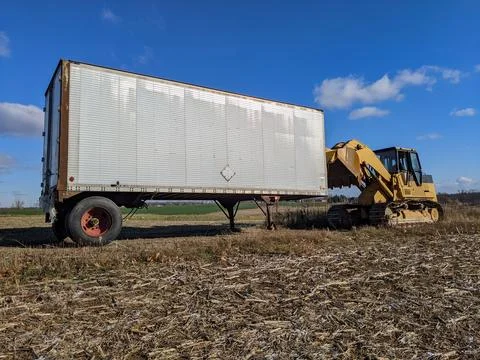 A Yellow Excavator Pulls an Abandoned Trailer in a Rural Field Under Blue Skies Stock Photos