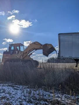 A Yellow Excavator Pulls an Abandoned Trailer in a Rural Field Under Blue Skies Stock-Fotos