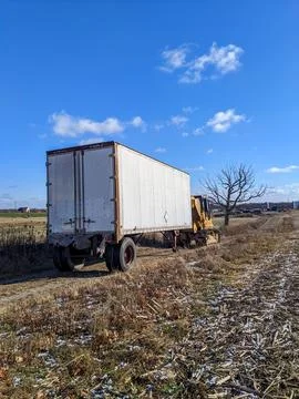 A Yellow Excavator Pulls an Abandoned Trailer in a Rural Field Under Blue Skies Stock Photos