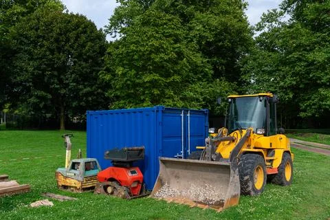 A yellow loader and red machine sit beside a blue storage container on green Stock Photos
