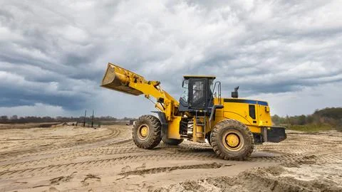 A yellow loader operates on sandy terrain under a gray sky, preparing the sit Stock-Fotos