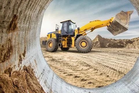 A yellow loader is seen through a concrete pipe at a construction site, with  스톡 사진