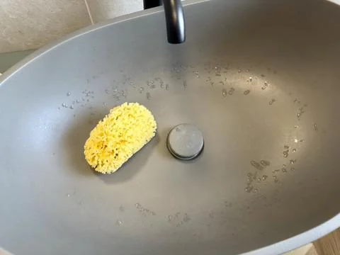 A yellow natural sponge sits inside a wet dark grey sink bowl  Stock Photos