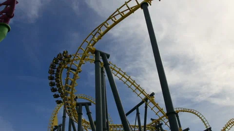 A yellow roller coaster twists and loops against a blue sky, with riders Stock Footage 287223809