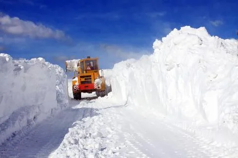 A yellow snowplow clears a path through high snowdrifts under a bright blue sky. Foto stock