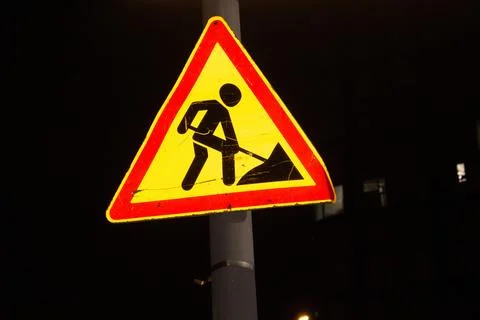Yellow triangular road sign with worker silhouette on black background at night Stock Photos