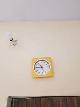 Yellow wall clock under a light bulb in a plain room Stock Photos