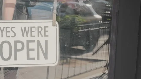 Yes We're Open sign hanging on a glass door. An employee opens the store Stock Footage 346771316