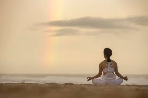 Yoga lotus pose meditation practice of young female on a beach with rainbow Stock Photos