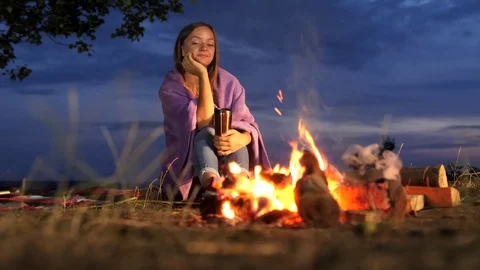 Yong woman sit next to a campfire with warm drinks and watch on campfire. Stock Footage