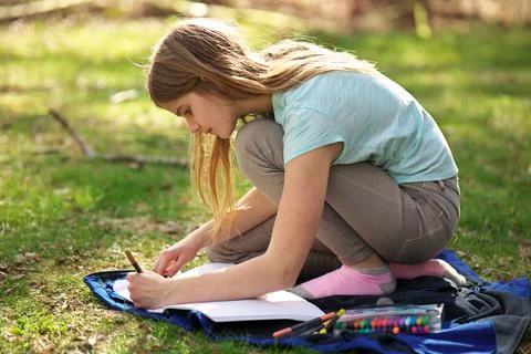 A young adolescent girl writes or colors in a notebook or journal on a blanket Stock Photos