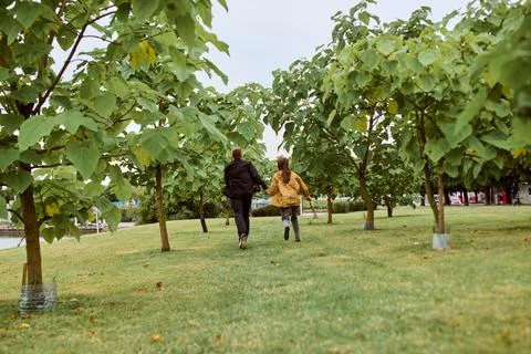 Young adults running between rows of lush green trees in park Stock Photos