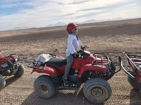 Young adventurer in a red cap confidently rides a red quad bike across the su Stock Photos