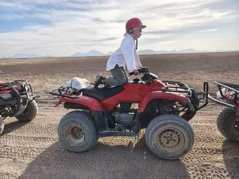 Young adventurer in a red cap confidently rides a red quad bike across the su Stock Photos