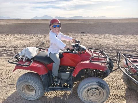 Young adventurer in a red cap confidently rides a red quad bike across the su Stock Photos