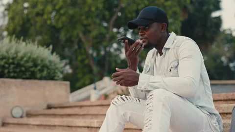 Young african american man using hands free mode on phone sitting in urban park. Stock Footage 304761316