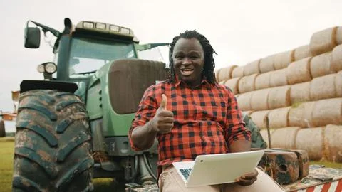 Young african farmer using tablet while sitting on green tractor. Haystack in Stock Photos