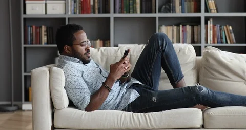 Young african guy using phone relax on sofa at home Stock Footage 122243552
