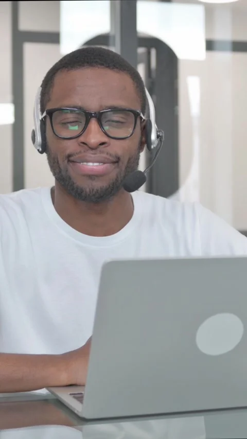 Young African Man with Headset Smiling at Camera while Working on Laptop, Stock Footage 324690295