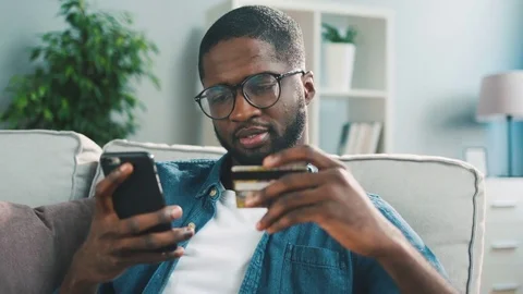 Young african man sitting on the couch, using smart phone for online shopping Stock Footage 75725169