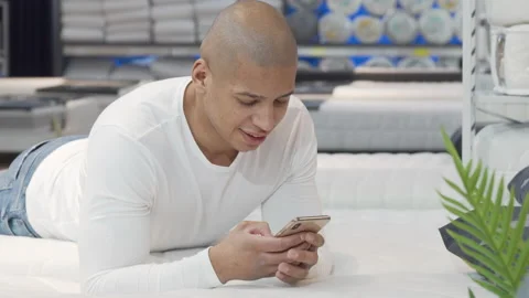 Young African man using smart phone while trying new bed at furniture store Stock Footage 148344856