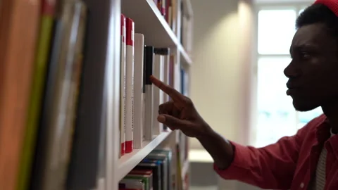 Young African millennial guy student picking up right book from bookshelf in Stock Footage 221674816