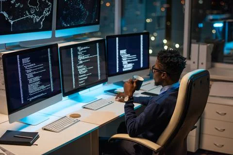 Young African programmer with drink looking at coded data on computer screen Foto stock