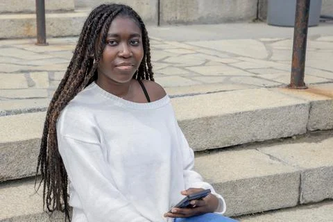 Young African student sitting on steps with smartphone, urban campus 库存照片