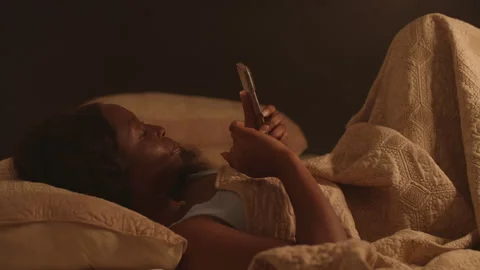 Young African woman using smartphone at night in bed, addicted to social media Видео 346077005